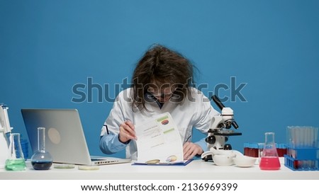 Goofy mad scientist using laptop and documents on camera, doing insane silly facial expressions and acting crazy. Foolish funny woman with messy wacky hair after smoke explosion.