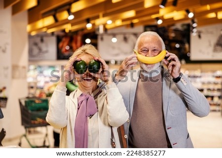 A goofy old couple is playing silly faces with fruits at supermarket.