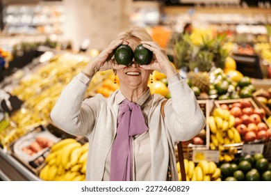 A goofy senior woman is playing with avocado and making silly faces at the hypermarket at fruit department. - Shutterstock ID 2272460829