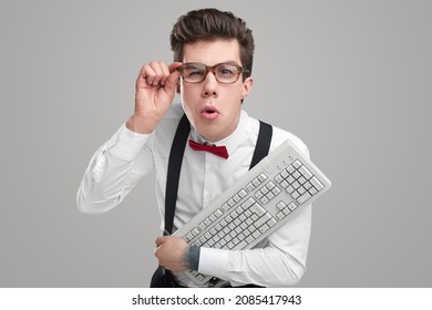 Goofy Young Man With Computer Keyboard Looking And Camera And Adjusting Glasses While Working Against Gray Background