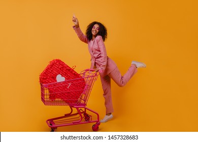 Gorgeous african girl in pink jacket enjoying shopping. Wonderful female shopaholic dancing on yellow background.