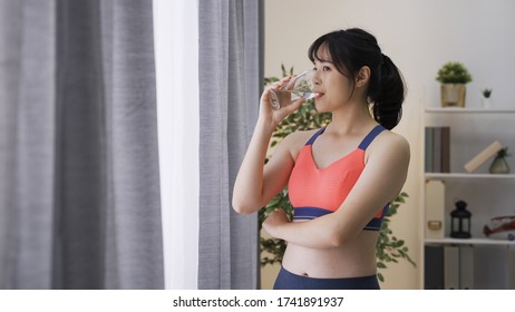 Gorgeous Asian Lady Standing With Single Arm Crossed Over Chest Feels Refreshed After Workout. Chinese Female Fitness Guru Looking Scenery Outside Of Window Is Enjoying Her Water And Sunny Morning