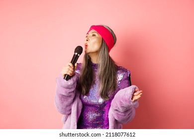 Gorgeous Asian Senior Woman Singing Karaoke In Mic, Perform Song And Looking Happy, Standing Over Pink Background