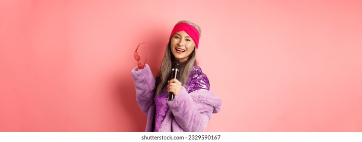 Gorgeous asian senior woman singing karaoke in mic, perform song and looking happy, standing over pink background. - Powered by Shutterstock