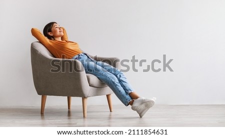 Gorgeous Asian woman sitting in comfy armchair, being lazy, feeling peaceful against white studio wall, panorama with copy space. Attractive young lady resting and relaxing with hands behind head