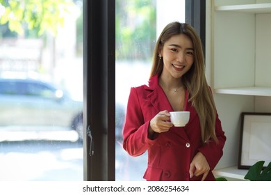 Gorgeous Asian Young Businesswoman In Red Suit Sipping Hot Coffee At Her Modern Office.