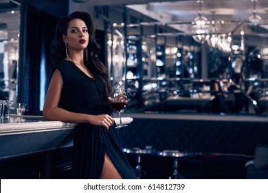 Gorgeous Beauty Young Brunette Woman In Dark Dress With Glass Of Wine Standing At The Bar In Luxury Interior