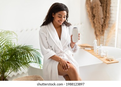 Gorgeous Black Lady Holding Shower Gel Bottle Advertising Cosmetic Product Sitting On Bathtub In Modern Bathroom At Home, Wearing White Bathrobe. Body Care Cosmetics Concept