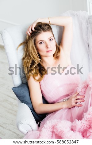 Gorgeous blond hair woman sitting in a vintage chair in a long pale pink ball dress. Elegant femininity. Classic white interior