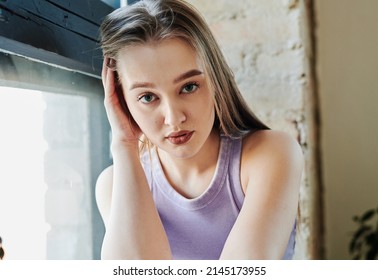 Gorgeous blond teenage girl in violet tanktop looking at camera in modern loft studio while sitting by window against wall