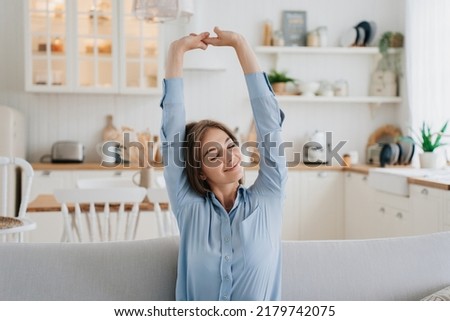 Gorgeous blonde caucasian young woman in blue shirt sitting eyes closed on couch stretching  rising up hands smiling. Pretty swedish girl  relaxing at home against kitchen. Comfort and cozy home.