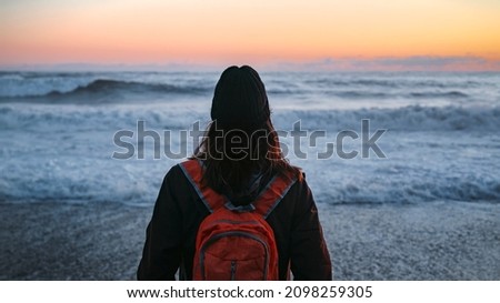 Gorgeous Brazilian young woman with an orange backpack looking out at the tranquil pacific ocean at Ruby Beach in Forks, Washington during a vivid colorful sunset on a warm summer evening