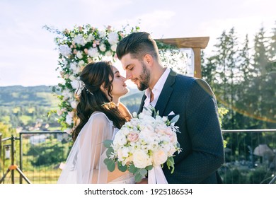 The gorgeous bride and groom stand right in front of the wedding altar outside. Girl holding a bouquet of flowers.