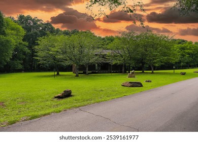 A gorgeous brown log cabin in the park surrounded by lush green grass and trees with red sky and powerful clouds at McIntosh Reserve Park in Whitesburg Georgia USA	 - Powered by Shutterstock