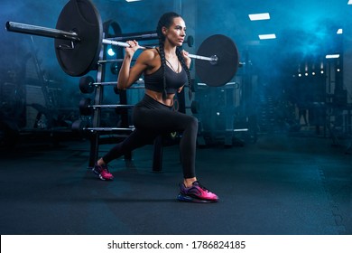 Gorgeous brunette female with long braids doing squats using barbell. Side view of srtong concentrated fitnesswoman with perfect muscular body training legs in gym in dark atmosphere and smoke.