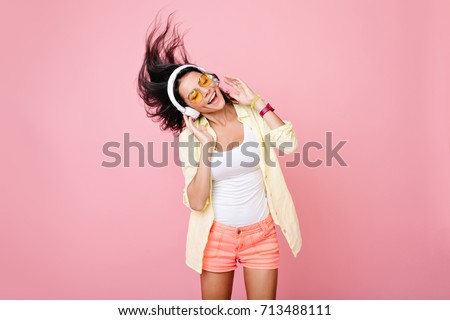 Gorgeous brunette lady in yellow glasses listening music in headphones and singing on pink background. Charming girl in shorts and earphones dancing with hair waving and eyes closed.