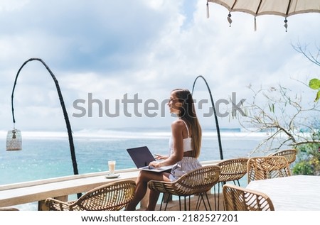 Gorgeous carefree young woman freelancer work online on laptop computer and enjoying sea view on vacations holidays. Smiling female digital nomad browsing internet on netbook while travel to Bali