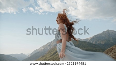 Gorgeous caucasian girl wearing a light dress is posing for a photo shoot on scenic mountain background while wind is blowing her red hair 