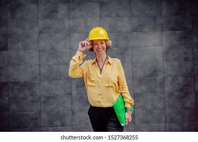 Gorgeous Caucasian Smiling Senior Female Architect With Helmet On Head And Documents Under Armpit Posing In Front Of Gray Wall Outdoors.