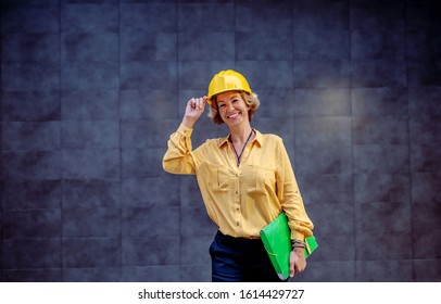 Gorgeous Caucasian Smiling Senior Female Architect With Helmet On Head And Documents Under Armpit Posing In Front Of Gray Wall Outdoors.