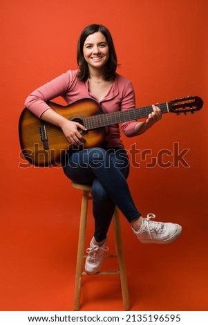 Gorgeous caucasian woman sitting on a stool and learning to play the acoustic guitar against an orange background