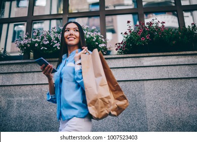 Gorgeous Cheerful Brunette Asian Woman Looking Away While Strolling At Spring Street With Shopping Bags And Modern Smartphone.Successful Female Person Enjoying Leisure Time Outdoors During City Walk