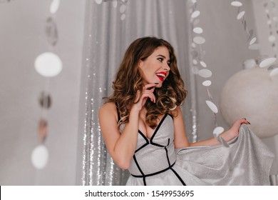 Gorgeous, Cheerful Long-haired Woman Rejoices At Holiday At New Year's Party. Portrait Of Girl Playing Her Brilliant Silver Dress On Background With Glitter