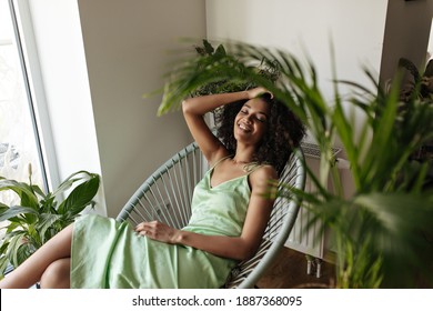 Gorgeous Curly Brunette Woman In Green Dress Sits On Armchair At Home. Dark-skinned Lady In Silk Stylish Outfit Smiles Sincerely.