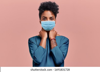 Gorgeous Dark Skinned Young Female With Afro Hairstyle Wearing Protective Mask Against COVID Virus, Tired Of Stress And Tension, Looks Confidently At The Camera, Poses Against Beige Studio Background