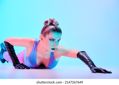 A gorgeous female model in bright extravagant clothes, leather gloves and high-heeled boots poses on the floor. Light blue background. Black makeup and avant-garde hairstyle. Vanguard fashion. 