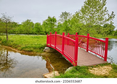 Gorgeous fire engine red walking bridge onto green island over stream leading to pond and geese