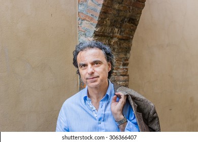 Gorgeous Forty Years Old Man With Piercing Green Eyes, Gray Hair And Three-day Macho  Beard Holds His Jacket Over His Shoulder Under An Ancient Brick Arch