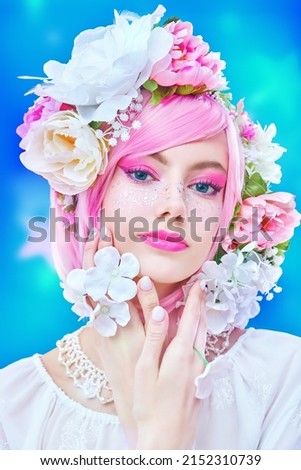Gorgeous girl with bright pink make-up poses in colored pink wig and flower wreath on head. Blue studio background. Beauty, makeup and hairstyle. 
