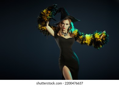 A Gorgeous Girl In A Witch's Hat, Black Dress And Feather Boa Poses On A Dark Background. Copy Space. Party Poster. Halloween Party. 