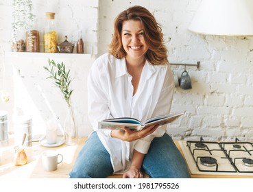 Gorgeous Happy Young Woman Plus Size Body Positive In Blue Jeans And White Shirt Reading Cooking Book In Home Kitchen