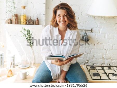Gorgeous happy young woman plus size body positive in blue jeans and white shirt reading cooking book in home kitchen