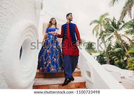Gorgeous Hindu groom in blue and red sherwani and bride in blue lehenga pose on the stairs in white house with greenery