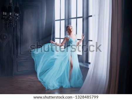 gorgeous image of graduate in 2019, girl in long blue gentle flying dress with bare leg stands alone, fabulous princess. elegant lady with blond hair in sunlight rays. Loft style interior background