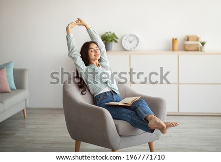 Gorgeous Indian woman stretching in comfy armchair while reading indoors. Attractive young lady with open book, enjoying lazy morning, having peaceful weekend. Stay home hobbies 商業照片 © 