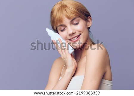 A gorgeous, joyful woman in a white T-shirt with fresh, well-groomed skin, with red wavy hair gathered in a tail stands on a lilac background with spray.Horizontal studio shot.