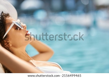 Gorgeous lady sunbathing while chilling out on lounger at hot day. Side view of relaxed girl in sunglasses, with both hands behind head, lying on sunbed next to swimming pool. Concept of chilling out.