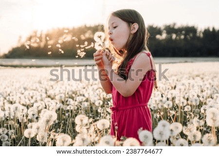 Gorgeous little girl in pink jumpsuit standing in meadow surrounded by white dandelions and blowing flowers. Female child playing with blossoms, seeds of blooming flying through air at sunset.