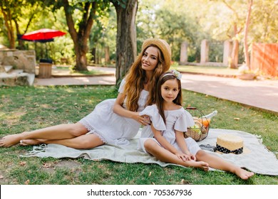 Gorgeous long-haired woman in straw hat and white dress have picnic with daughter in good summer day. Outdoor portrait of pretty little girl spending time with mother in park. - Powered by Shutterstock