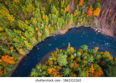 Gorgeous look down travel aerial of red dirt cliffs and fall foliage leaves on deciduous trees and evergreen trees lining the curve of a river bend on the Bad River in Mellen Wisconsin.