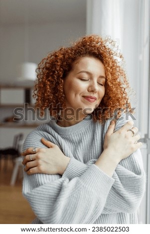 Gorgeous lovely charming redhead female with cute curls standing with closed eyes next to window at home, hugging herself, touching her favorite soft blue sweater, enjoying comfort of fabric