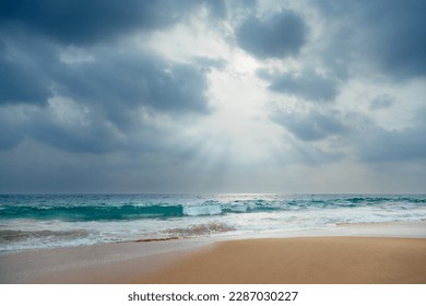 A gorgeous low-angle view of a sandy ocean beach with turquoise waves under a dramatic cloudy sky with sunlight through it. - Powered by Shutterstock