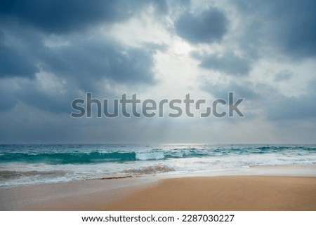 A gorgeous low-angle view of a sandy ocean beach with turquoise waves under a dramatic cloudy sky with sunlight through it.