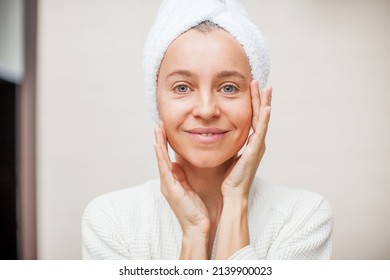 Gorgeous Mid Age Adult 40 Years Woman Standing In Bathroom After Shower Touching Face, Looking At Camera Doing Morning Beauty Routine. Older Dry Skin Care Concept.