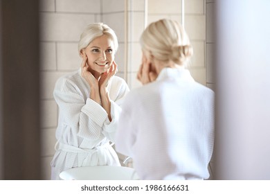 Gorgeous mid age adult 50 years old blonde woman standing in bathroom wearing bathrobe touching face, looking at reflection in mirror smiling doing morning beauty routine. Antiage skin care concept. - Powered by Shutterstock