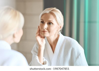 Gorgeous mid age adult 50 years old blonde woman standing in bathroom after shower touching face, looking at reflection in mirror doing morning beauty routine. Older dry skin care concept. - Powered by Shutterstock